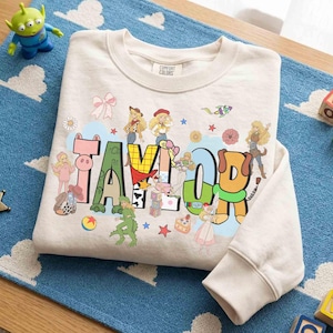 May include: A beige long-sleeve sweatshirt featuring the name TAYLOR in large, colorful, patterned block letters. The design includes various cartoon characters, a rocket, stars, flowers, and a bow, with a Comfort Colors label at the neck.