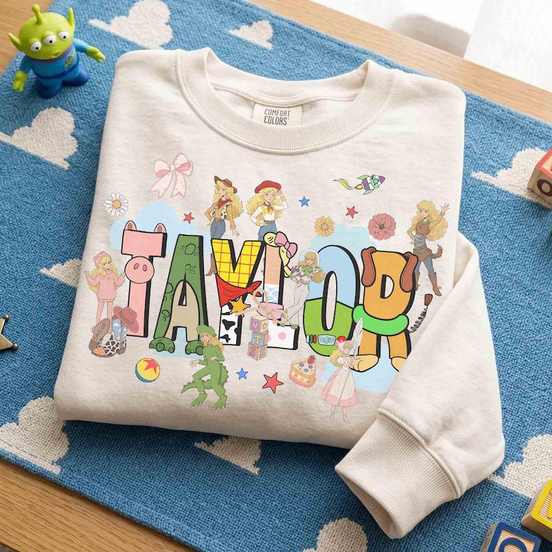May include: A beige long-sleeve sweatshirt featuring the name TAYLOR in large, colorful, patterned block letters. The design includes various cartoon characters, a rocket, stars, flowers, and a bow, with a Comfort Colors label at the neck.