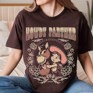 Puede incluir: Una camiseta de manga corta de color marr&oacute;n oscuro con el texto HOWDY PARTNER en una fuente degradada. El gr&aacute;fico muestra a una vaquera de dibujos animados abrazando a un caballo, rodeada por una corona floral y una cuerda, con PIXAR abajo.