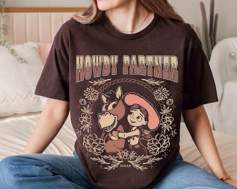 Puede incluir: Una camiseta de manga corta de color marr&oacute;n oscuro con el texto HOWDY PARTNER en una fuente degradada. El gr&aacute;fico muestra a una vaquera de dibujos animados abrazando a un caballo, rodeada por una corona floral y una cuerda, con PIXAR abajo.