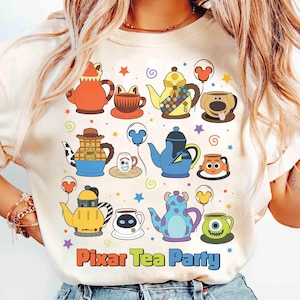 May include: Cream-colored t-shirt featuring a colorful "Pixar Tea Party" design. The design includes various cartoon teapots and teacups with character-inspired designs, surrounded by stars and swirls. The text "Pixar Tea Party" is at the bottom.