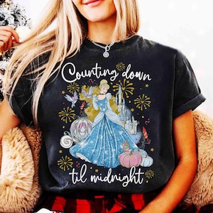 Può includere: T-shirt nera con Cenerentola in abito blu, una carrozza a forma di zucca e fuochi d'artificio. Il testo "Counting down til midnight" è in scrittura bianca. Il design ha un tema fiabesco.