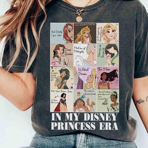 May include: Dark gray t-shirt featuring a collage of Disney Princess illustrations with quotes. The shirt includes the text "IN MY DISNEY PRINCESS ERA" in a white, bold font. The shirt is a casual wear item.