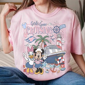 Peut inclure: T-shirt rose clair avec l'inscription "Girls Gone Cruising" et un motif repr&eacute;sentant Mickey et Daisy Duck, un bateau de croisi&egrave;re, un phare et des palmiers. Le motif comprend un billet de croisi&egrave;re et des feux d'artifice.