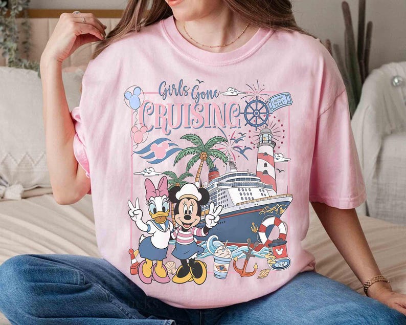 Peut inclure: T-shirt rose clair avec l'inscription "Girls Gone Cruising" et un motif repr&eacute;sentant Mickey et Daisy Duck, un bateau de croisi&egrave;re, un phare et des palmiers. Le motif comprend un billet de croisi&egrave;re et des feux d'artifice.