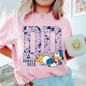 May include: A light pink t-shirt featuring a navy blue "DD" design filled with Donald Duck illustrations. Below the design is the text "DONALD DUCK" and a cartoon image of Donald Duck. A white mug with a smiley face is also visible.
