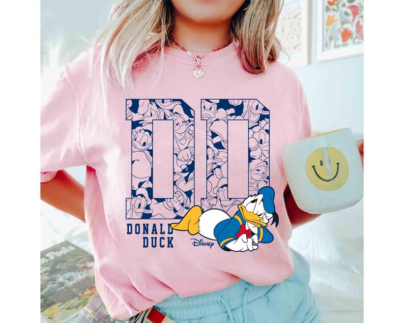 May include: A light pink t-shirt featuring a navy blue "DD" design filled with Donald Duck illustrations. Below the design is the text "DONALD DUCK" and a cartoon image of Donald Duck. A white mug with a smiley face is also visible.
