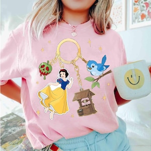 May include: A light pink t-shirt featuring a gold keychain with Snow White and other fairytale-themed charms. The charms include a red apple, a blue bird, and a wishing well. A white mug with a smiley face is held in the image.