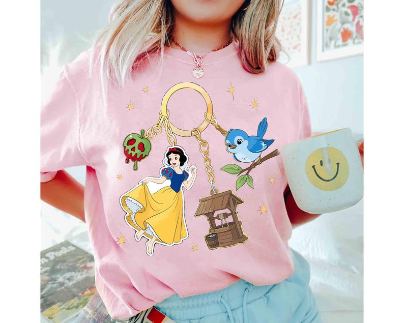 May include: A light pink t-shirt featuring a gold keychain with Snow White and other fairytale-themed charms. The charms include a red apple, a blue bird, and a wishing well. A white mug with a smiley face is held in the image.