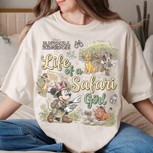 May include: Cream-colored t-shirt featuring a graphic design with Disney's Animal Kingdom theme. The design includes the text "Life of a Safari Girl" and illustrations of Simba, Timon, Pumbaa, and Minnie Mouse in safari attire.