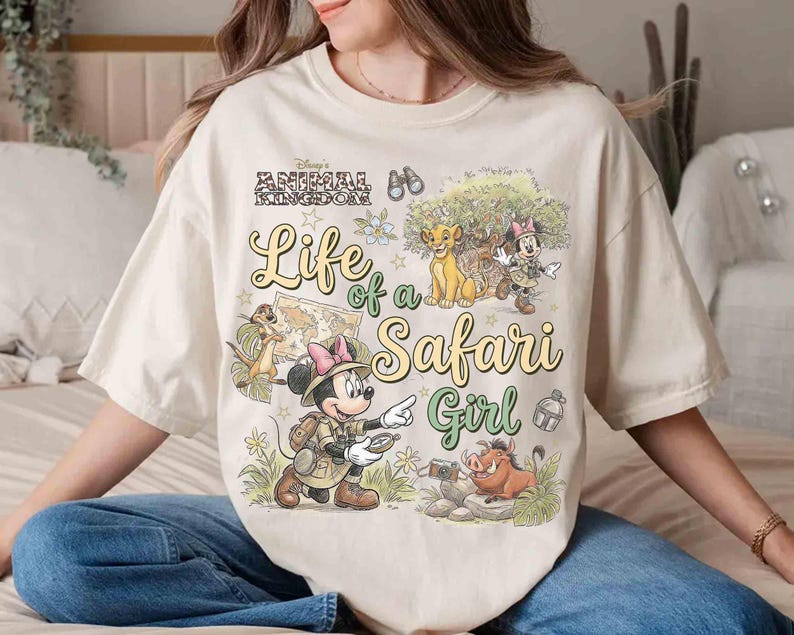 May include: Cream-colored t-shirt featuring a graphic design with Disney's Animal Kingdom theme. The design includes the text "Life of a Safari Girl" and illustrations of Simba, Timon, Pumbaa, and Minnie Mouse in safari attire.