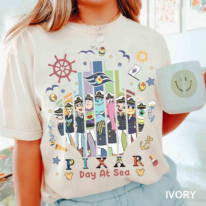 K&ouml;nnte beinhalten: Elfenbeinfarbenes T-Shirt mit einem farbenfrohen Aufdruck von Pixar-Charakteren in Matrosenuniformen. Das Design enth&auml;lt den Text "PIXAR Day At Sea" und nautische Elemente wie ein Steuerrad und einen Anker. Eine wei&szlig;e Tasse mit einem Smiley ist sichtbar.
