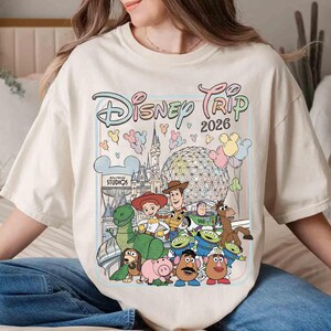 May include: Cream-colored t-shirt featuring a colorful graphic of Disney characters and the text "Disney Trip 2026." The design includes iconic landmarks and characters from various Disney properties, such as Toy Story and Hollywood Studios.