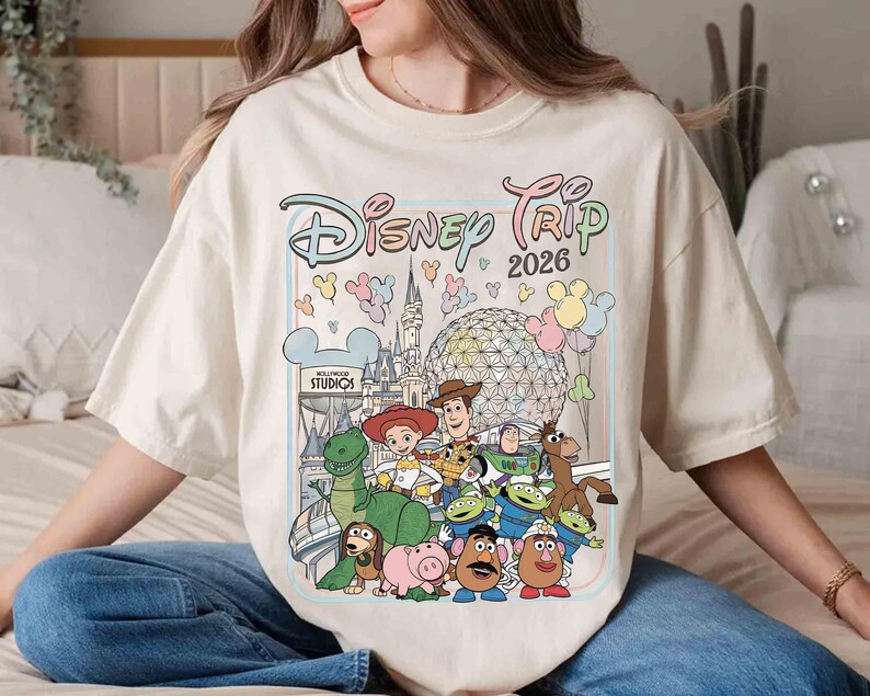 May include: Cream-colored t-shirt featuring a colorful graphic of Disney characters and the text "Disney Trip 2026." The design includes iconic landmarks and characters from various Disney properties, such as Toy Story and Hollywood Studios.