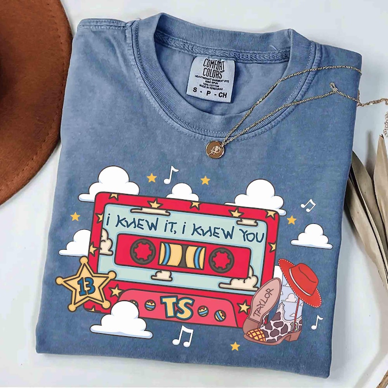 May include: A blue crew neck t-shirt featuring a graphic of a red cassette tape with the text "I KNEW IT, I KNEW YOU" and "TS". The design includes a yellow star with the number 13, white clouds, musical notes, and a cowboy boot with a red hat.
