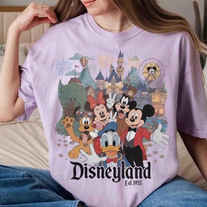 May include: A light purple t-shirt featuring a colorful graphic of Disneyland characters including Mickey Mouse, Minnie Mouse, Donald Duck, Goofy, and Pluto. The Disneyland logo and "Est. 1955" are printed below the characters.