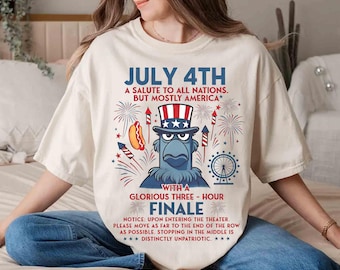 以下が含まれることがあります： 7月4日の愛国的なデザインのクリーム色のTシャツ。グラフィックには、アンクルサムの帽子をかぶった漫画のワシ、花火、ホットドッグ、そして「JULY 4TH」の文字が含まれています。シャツには追加のテキストも含まれています。