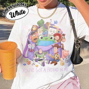 May include: A white t-shirt featuring a graphic print of various Toy Story characters emerging from a purple backpack. The design includes Woody, Buzz Lightyear, and others, with the text "YOU'VE GOT A FRIEND IN ME" printed in purple below the backpack.