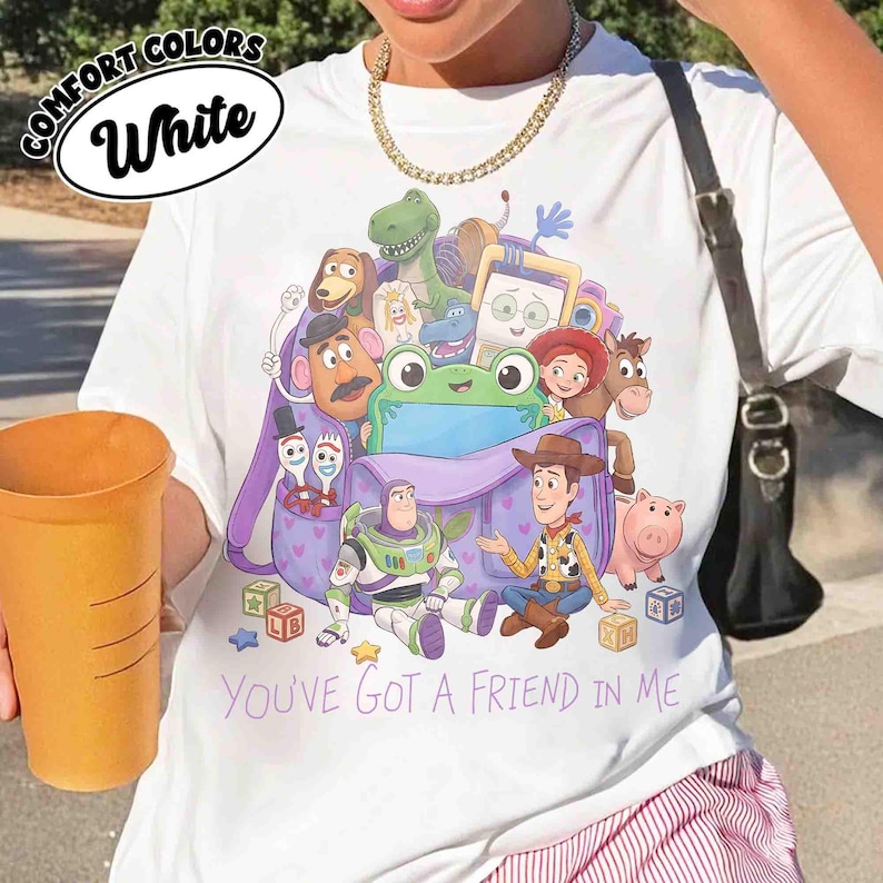 May include: A white t-shirt featuring a graphic print of various Toy Story characters emerging from a purple backpack. The design includes Woody, Buzz Lightyear, and others, with the text "YOU'VE GOT A FRIEND IN ME" printed in purple below the backpack.