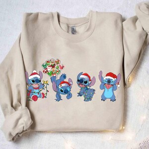 Puede incluir: Sudadera beige con cuatro personajes de dibujos animados azules, cada uno con un gorro de Pap&aacute; Noel. Los personajes est&aacute;n en varias poses, algunos sosteniendo luces navide&ntilde;as y adornos. Un dise&ntilde;o festivo y colorido para la temporada navide&ntilde;a.
