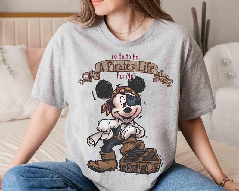 Puede incluir: Una camiseta gris con una imagen de dibujos animados de Mickey Mouse vestido de pirata. El texto "Yo Ho, Yo Ho, A Pirates Life For Me!" est&aacute; impreso en la camiseta.