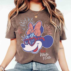 Op de afbeelding: Bruin t-shirt met een cartoonontwerp van Minnie Mouse. Het ontwerp omvat een rood-witte geruite strik, hartvormige zonnebrillen en vuurwerk. De tekst "White & Cute" is gedrukt.