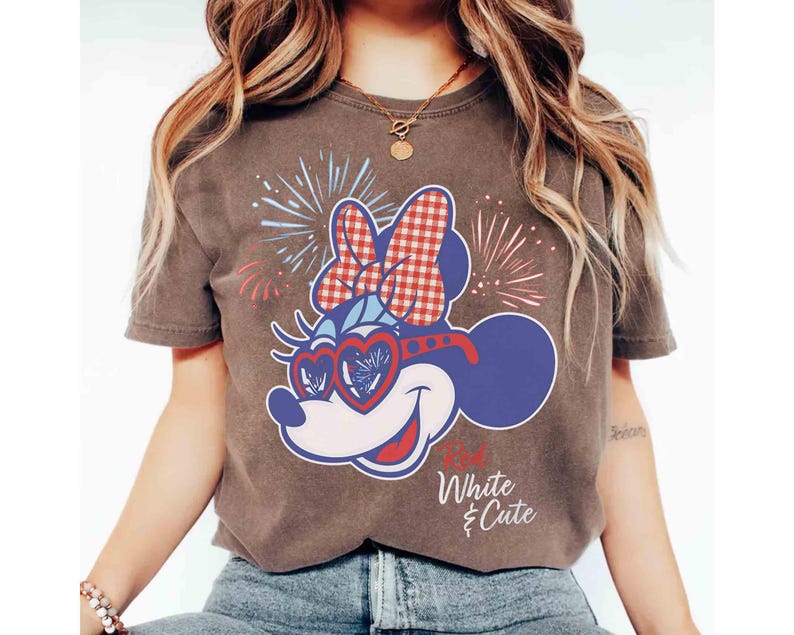 Op de afbeelding: Bruin t-shirt met een cartoonontwerp van Minnie Mouse. Het ontwerp omvat een rood-witte geruite strik, hartvormige zonnebrillen en vuurwerk. De tekst "White & Cute" is gedrukt.