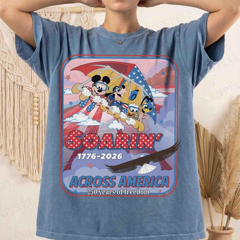 May include: Blue t-shirt featuring a graphic of Mickey Mouse, Donald Duck, and Goofy soaring in a hang glider. The design includes the text "Soarin'" in red and white stars, with "1776-2026" and "Across America" below, celebrating 250 years of freedom.