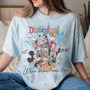 May include: Light blue t-shirt featuring a colorful Disneyland graphic with Mickey Mouse, Minnie Mouse, Donald Duck, Daisy Duck, Goofy, and Pluto. The shirt includes the text "Disneyland", "est. 1955", and "Where dreams come true."