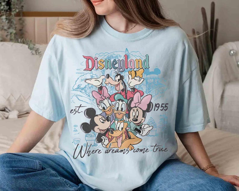 May include: Light blue t-shirt featuring a colorful Disneyland graphic with Mickey Mouse, Minnie Mouse, Donald Duck, Daisy Duck, Goofy, and Pluto. The shirt includes the text "Disneyland", "est. 1955", and "Where dreams come true."