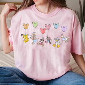 May include: A light pink t-shirt featuring a colorful graphic of Disney characters holding Mickey Mouse-shaped balloons. Pluto, Goofy, Mickey, Minnie, Donald, and Daisy are depicted. The balloons are in various pastel colors.