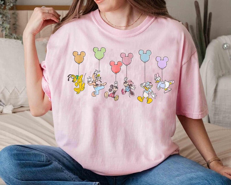 May include: A light pink t-shirt featuring a colorful graphic of Disney characters holding Mickey Mouse-shaped balloons. Pluto, Goofy, Mickey, Minnie, Donald, and Daisy are depicted. The balloons are in various pastel colors.