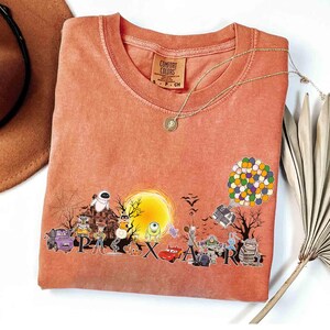 May include: Coral-colored Comfort Colors t-shirt featuring a graphic of Pixar characters in front of a full moon. The word "Pixar" is spelled out across the bottom. A gold necklace and a brown hat are also visible.