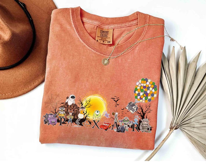 May include: Coral-colored Comfort Colors t-shirt featuring a graphic of Pixar characters in front of a full moon. The word "Pixar" is spelled out across the bottom. A gold necklace and a brown hat are also visible.