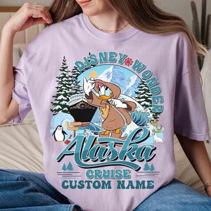 May include: A light purple t-shirt featuring a graphic of Donald Duck in a brown coat, with the text "Disney Wonder Alaska Cruise Custom Name". The design includes a cruise ship, mountains, and a penguin.