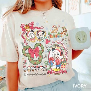 May include: Ivory-colored t-shirt featuring a festive Christmas design with Mickey Mouse, Minnie Mouse, Chip and Dale, and other Disney characters. The shirt includes the text "Very Merry Christmas" and "The most magical place on Earth."