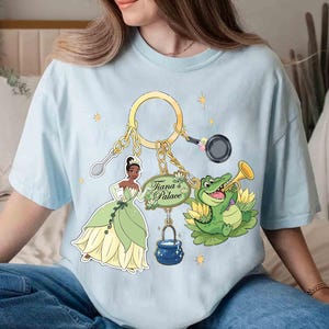 May include: Light blue t-shirt featuring a gold keychain with cartoon images of Tiana, a frog playing a trumpet, a spoon, a frying pan, and a pot. The text "Tiana's Palace" is on a green sign.