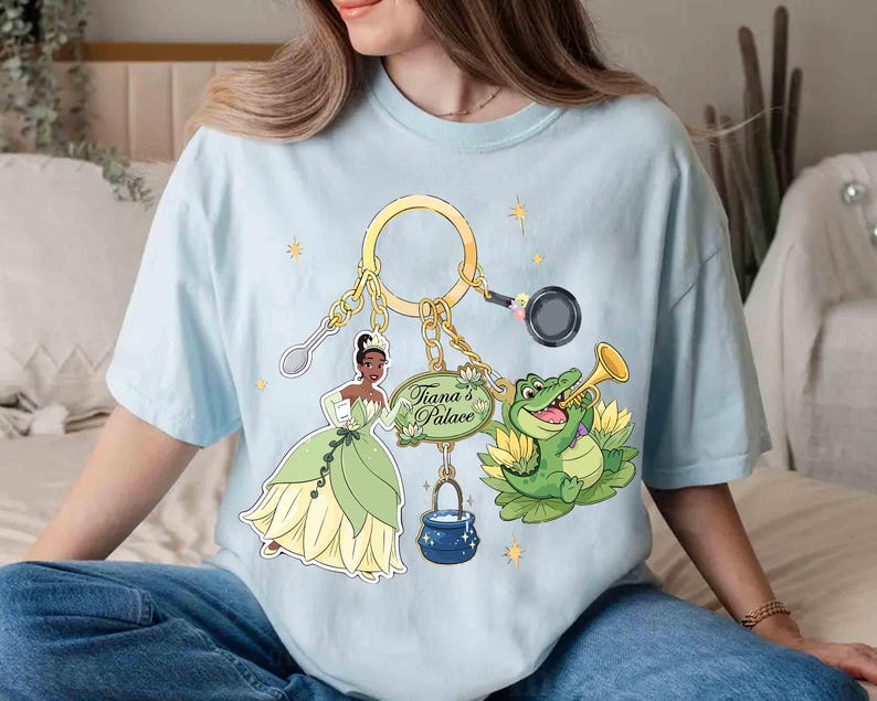 May include: Light blue t-shirt featuring a gold keychain with cartoon images of Tiana, a frog playing a trumpet, a spoon, a frying pan, and a pot. The text "Tiana's Palace" is on a green sign.