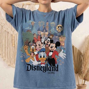 May include: Blue t-shirt featuring a colorful graphic of Disney characters including Mickey Mouse, Minnie Mouse, Donald Duck, and Pluto. The graphic includes a castle, fireworks, and the text "Disneyland Est.1955".