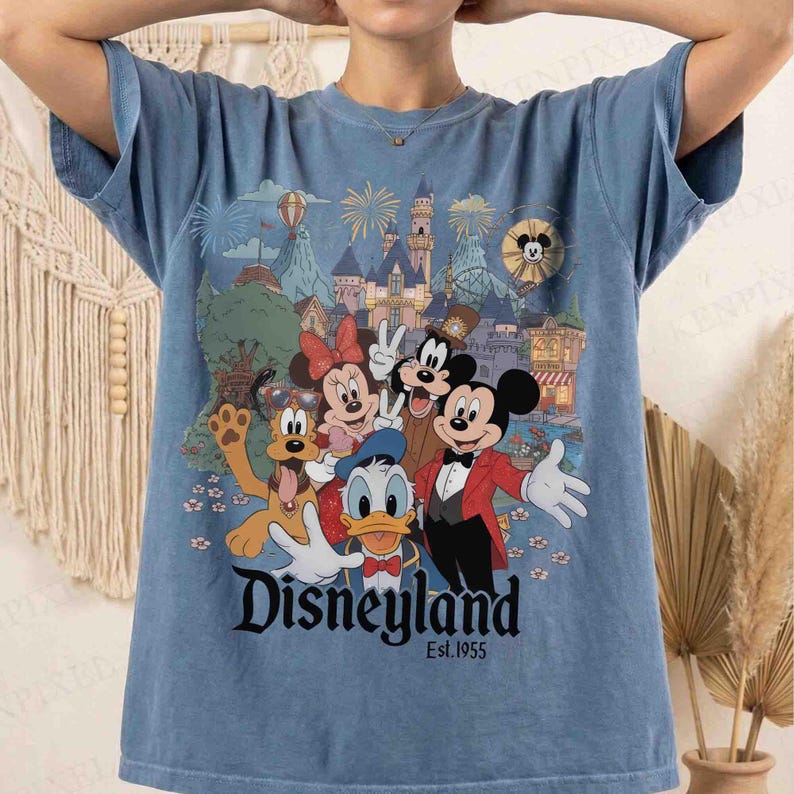 May include: Blue t-shirt featuring a colorful graphic of Disney characters including Mickey Mouse, Minnie Mouse, Donald Duck, and Pluto. The graphic includes a castle, fireworks, and the text "Disneyland Est.1955".