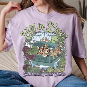 May include: A light purple t-shirt featuring a cartoon graphic of a boat with Disney characters and the text "Get in Loser, We're learning about Lettuce". The shirt has a crew neck and short sleeves.