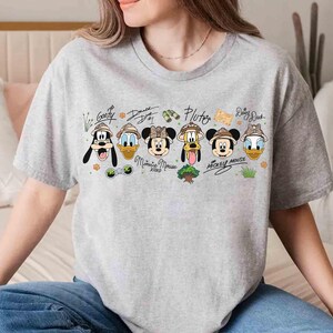 May include: A light gray t-shirt featuring cartoon images of Disney characters Goofy, Donald Duck, Minnie Mouse, Pluto, Mickey Mouse, and Daisy Duck, each wearing safari hats. The characters' names are written above them in a handwritten style.
