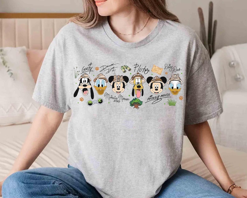 May include: A light gray t-shirt featuring cartoon images of Disney characters Goofy, Donald Duck, Minnie Mouse, Pluto, Mickey Mouse, and Daisy Duck, each wearing safari hats. The characters' names are written above them in a handwritten style.