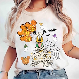 Könnte beinhalten: Cremefarbenes T-Shirt mit einem Halloween-Motiv. Die Grafik zeigt einen mumienverpackten Hund, Mickey Mouse Kürbisballons, Geister, Fledermäuse und ein Spinnennetz. Das Design ist in Orange, Schwarz, Grün und Weiß gehalten.