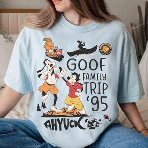 Pode incluir: Camiseta azul clara com um desenho animado do Pateta e Max. A camisa tem o texto "GOOF FAMILY TRIP '95" e "AHYUCK", juntamente com outros elementos de desenho animado. Uma camiseta divertida e casual.