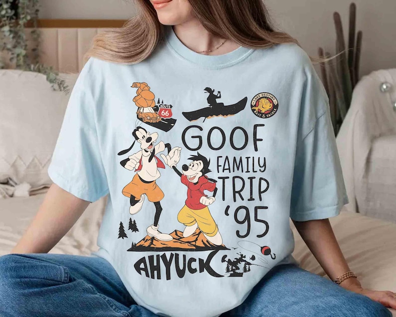 Pode incluir: Camiseta azul clara com um desenho animado do Pateta e Max. A camisa tem o texto "GOOF FAMILY TRIP '95" e "AHYUCK", juntamente com outros elementos de desenho animado. Uma camiseta divertida e casual.