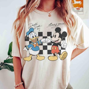 May include: Cream-colored t-shirt featuring a graphic of Donald Duck and Mickey Mouse fist-bumping. Donald Duck is wearing a blue sailor suit and hat. Mickey Mouse is wearing red shorts and yellow shoes. The text "Donald Duck #1" and "Mickey Mouse" are above the characters.