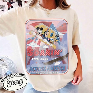 May include: Ivory-colored t-shirt featuring a graphic of Mickey Mouse, Donald Duck, and Goofy soaring with an American flag-themed parachute. The design includes the text "SOARIN' 1776-2026 ACROSS AMERICA 250 years of freedom".