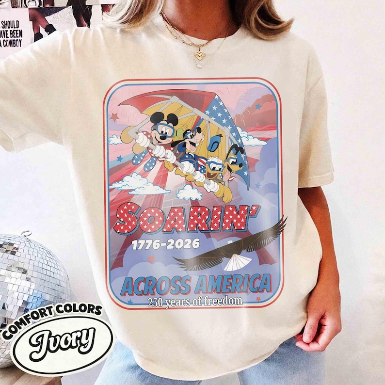 May include: Ivory-colored t-shirt featuring a graphic of Mickey Mouse, Donald Duck, and Goofy soaring with an American flag-themed parachute. The design includes the text "SOARIN' 1776-2026 ACROSS AMERICA 250 years of freedom".