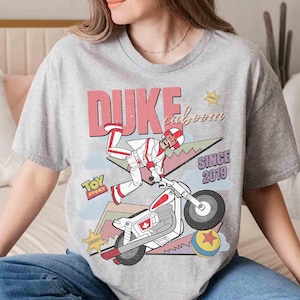 May include: A heather gray t-shirt featuring a graphic of Duke Caboom from Toy Story riding a motorcycle. The design includes the text "DUKE caboom" and "SINCE 2019". A sheriff badge and a Toy Story logo are also visible.